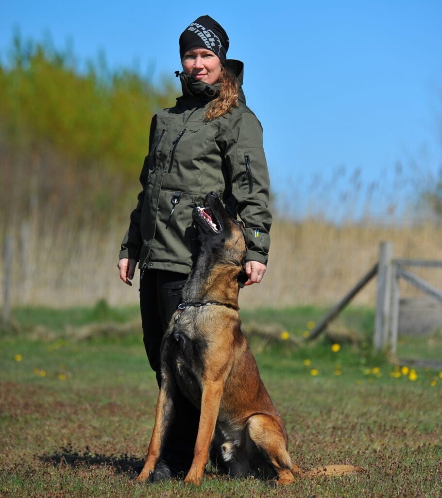 Person træner hund i grøn jakke, udendørs landskab.